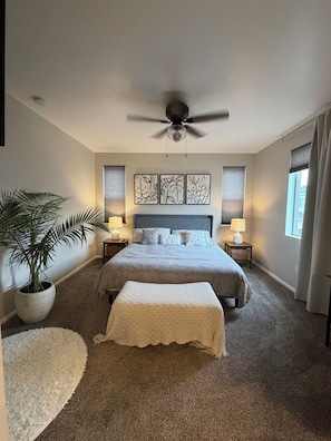 A bedroom with a large bed, a ceiling fan, a potted plant, and artwork on the wall.