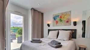 Ein Schlafzimmer mit einem Bett, zwei Kissen, einem Nachttisch, einer Lampe, einem Gemälde und Blick ins Freie.