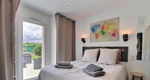 A bedroom with a bed, two pillows, a bedside table, a lamp, a painting, and a view of the outdoors. - L'écrin - PLOURHAN - Gîte (PLOURHAN)