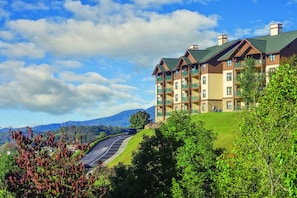 Un hotel de varios pisos con balcones, situado en una ladera con vistas a montañas y una carretera sinuosa.