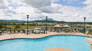 En swimmingpool med liggestole og parasoller, omgivet af et hegn, med udsigt til et bjerg og en by i baggrunden.