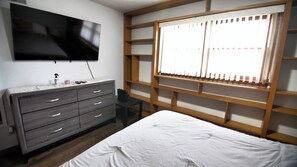 Ein Schlafzimmer mit einem Bett, einer Kommode, einem an der Wand montierten Fernseher und einem Fenster mit Jalousien.