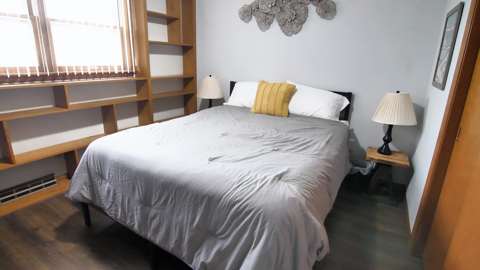 A bedroom with a bed, two bedside tables with lamps, a window with blinds, and a wall decoration.