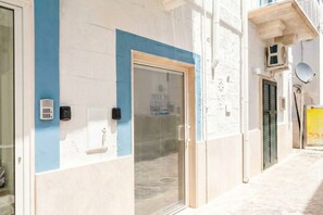 Une façade d’immeuble aux couleurs bleu et blanc, une porte en verre avec un cadre en métal et une petite fenêtre ornée d’une grille métallique.