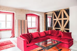 Un sofá seccional rojo, una mesa de centro de vidrio y una pared decorativa de madera en una habitación con ventanas amplias y una cortina roja.