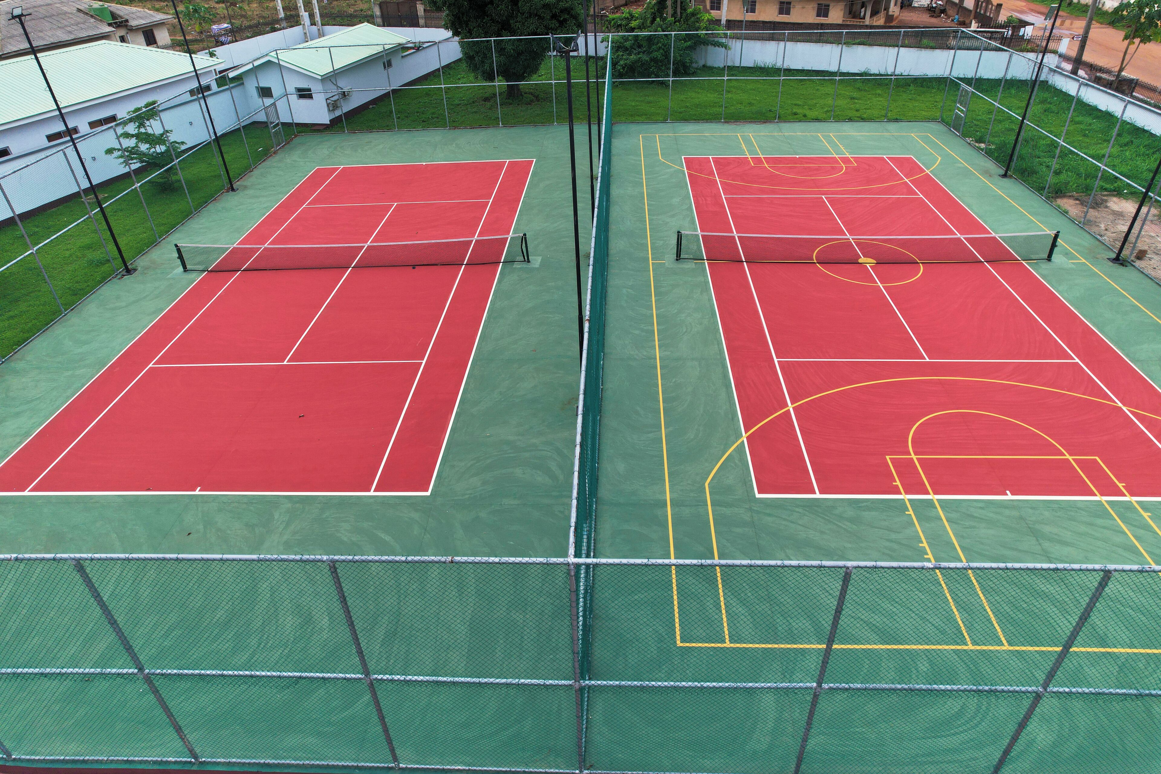 A tennis court with a net, surrounded by a fence, and adjacent buildings.