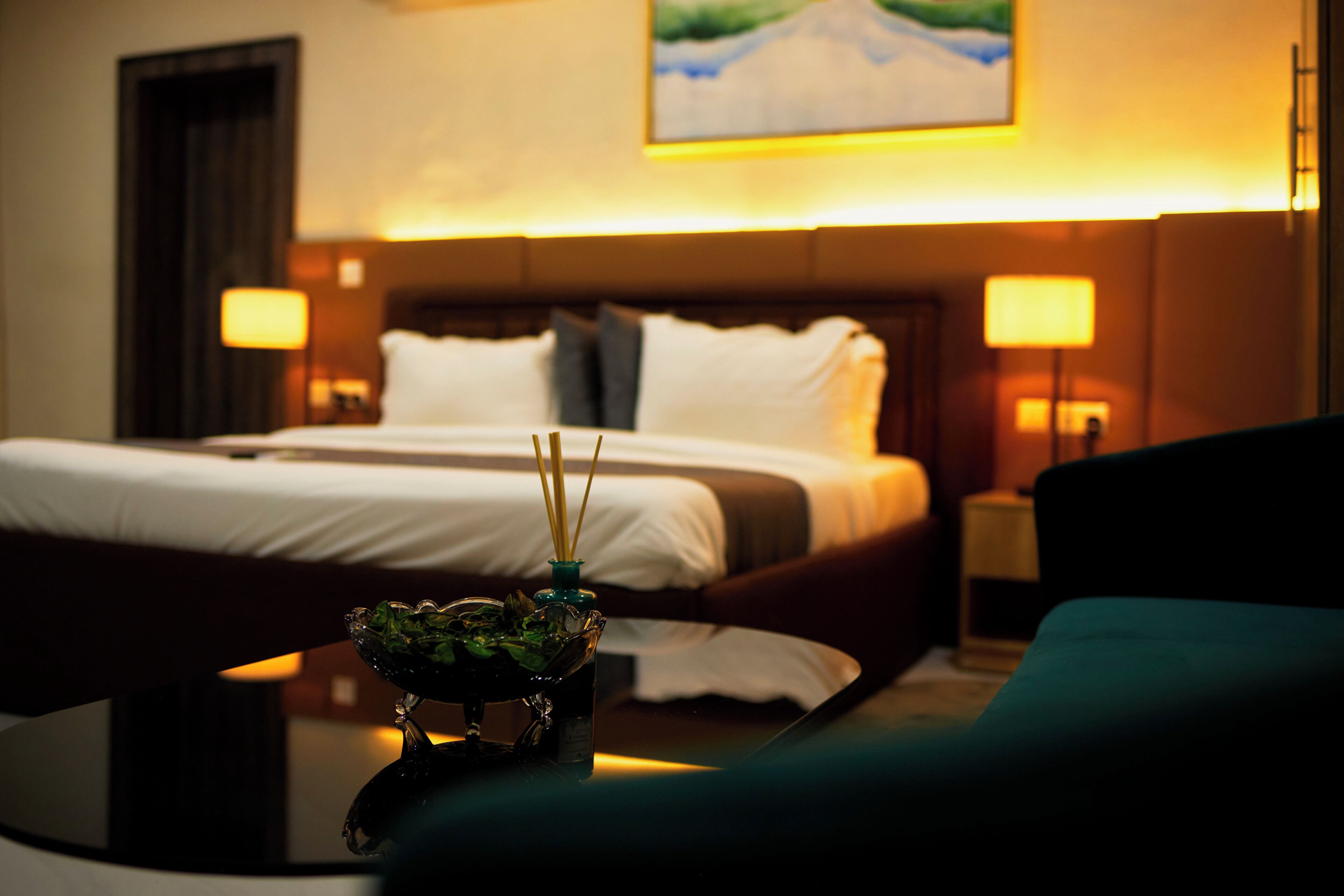 Habitación de hotel con cama, lámparas de noche, una mesita con un objeto decorativo y un cuadro en la pared.
