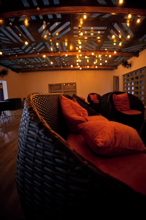 Una habitación con un sofá de mimbre, una silla negra, vigas a la vista en el techo y luces colgantes.