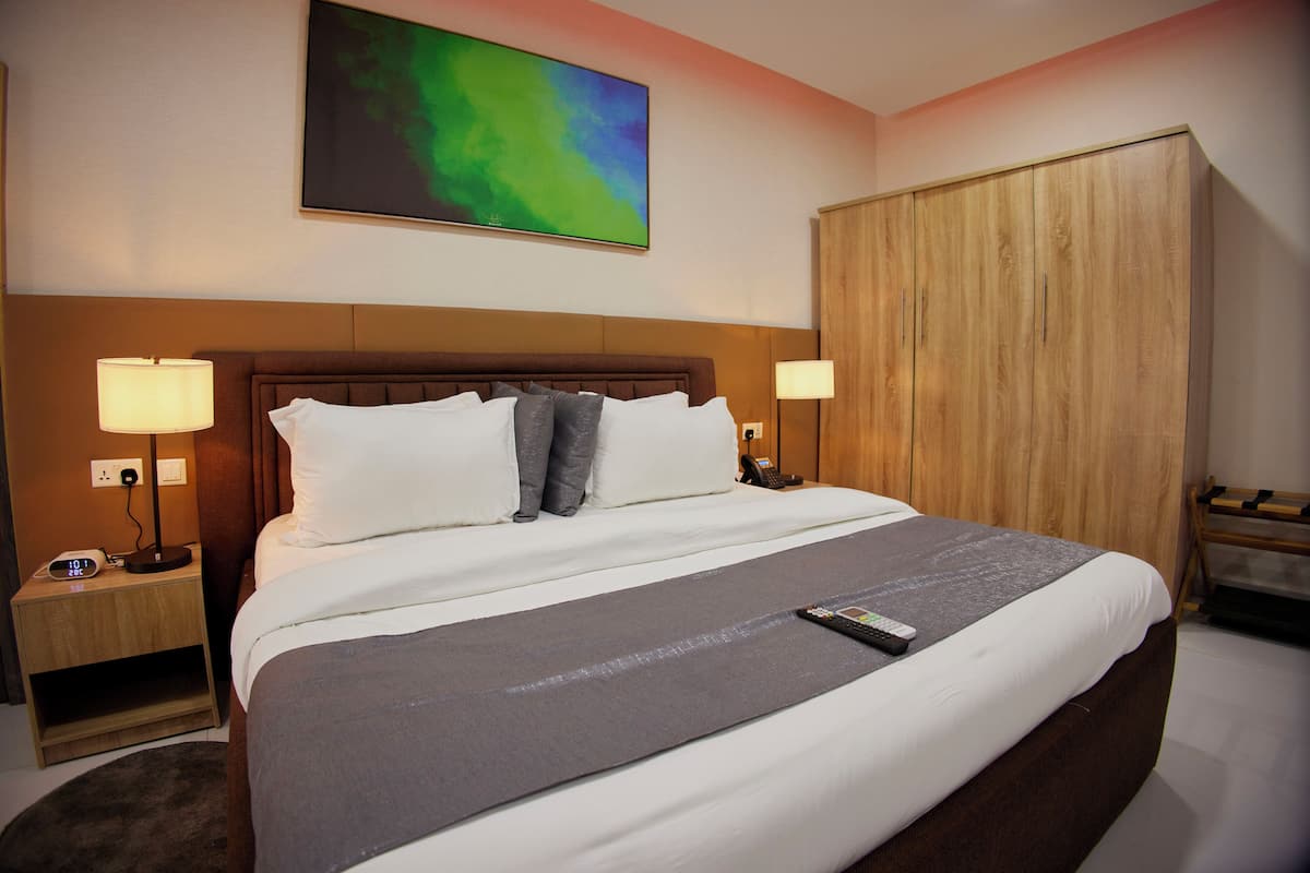Una habitación de hotel con una cama grande, dos mesitas de noche con lámparas, un armario de madera y un cuadro abstracto colorido en la pared.