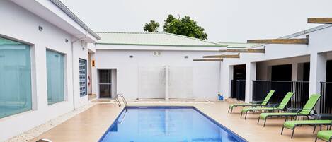 Ein Swimmingpool mit klarem, blauem Wasser, umgeben von Liegestühlen und einem Gebäude mit großen Fenstern.