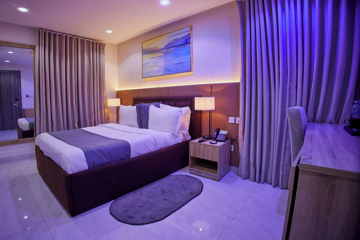 Una habitación de hotel moderna con una cama grande, lámparas de noche, un escritorio con silla, una mesita y un cuadro en la pared.