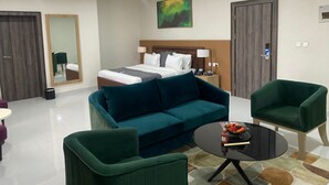 Una habitación de hotel moderna con cama, sofá verde, dos sillones, una mesita y un cuadro en la pared.