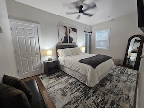 A bedroom with a bed, bedside lamps, a ceiling fan, a window with blinds, a door, and a mirror. - Charming 1-bedroom apartment with AC in Fort Mill (Fort Mill)