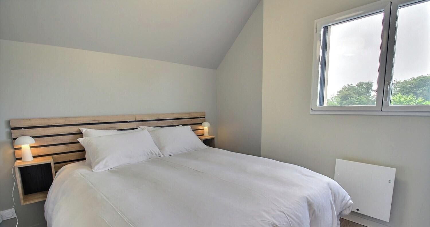 A bedroom with a slatted wooden headboard, a bed with white bedding, a bedside lamp, and a window with a view of greenery.