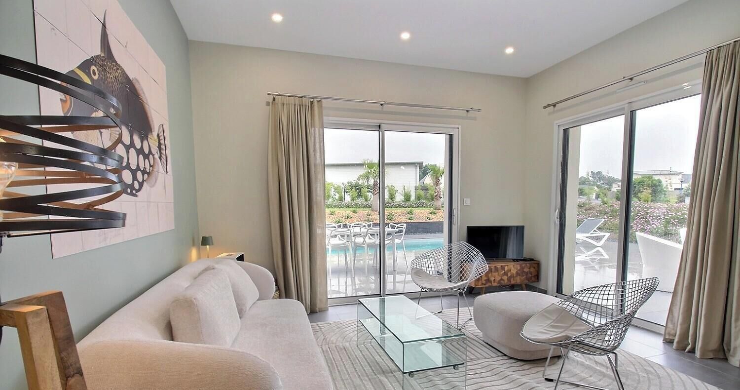 A modern living room with a sofa, a glass coffee table, and a TV. There is a large window with a view of a pool and outdoor furniture.