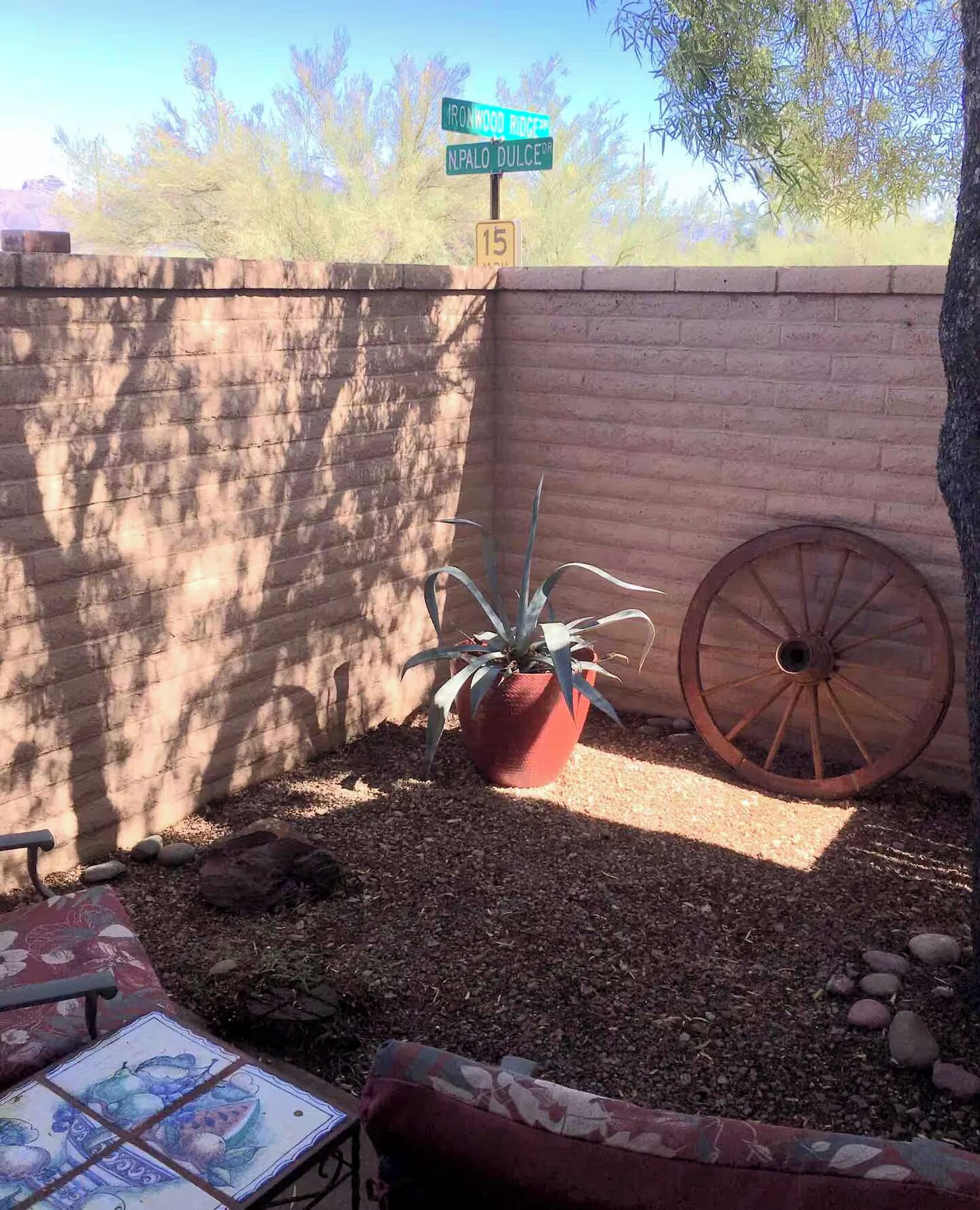 A brick wall with a street sign, a potted plant, a wooden wheel, and a tiled table.
