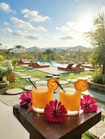 Escena junto a la piscina con dos bebidas sobre una mesa, rodeada de sillones de playa y sombrillas, con vistas a montañas al fondo.