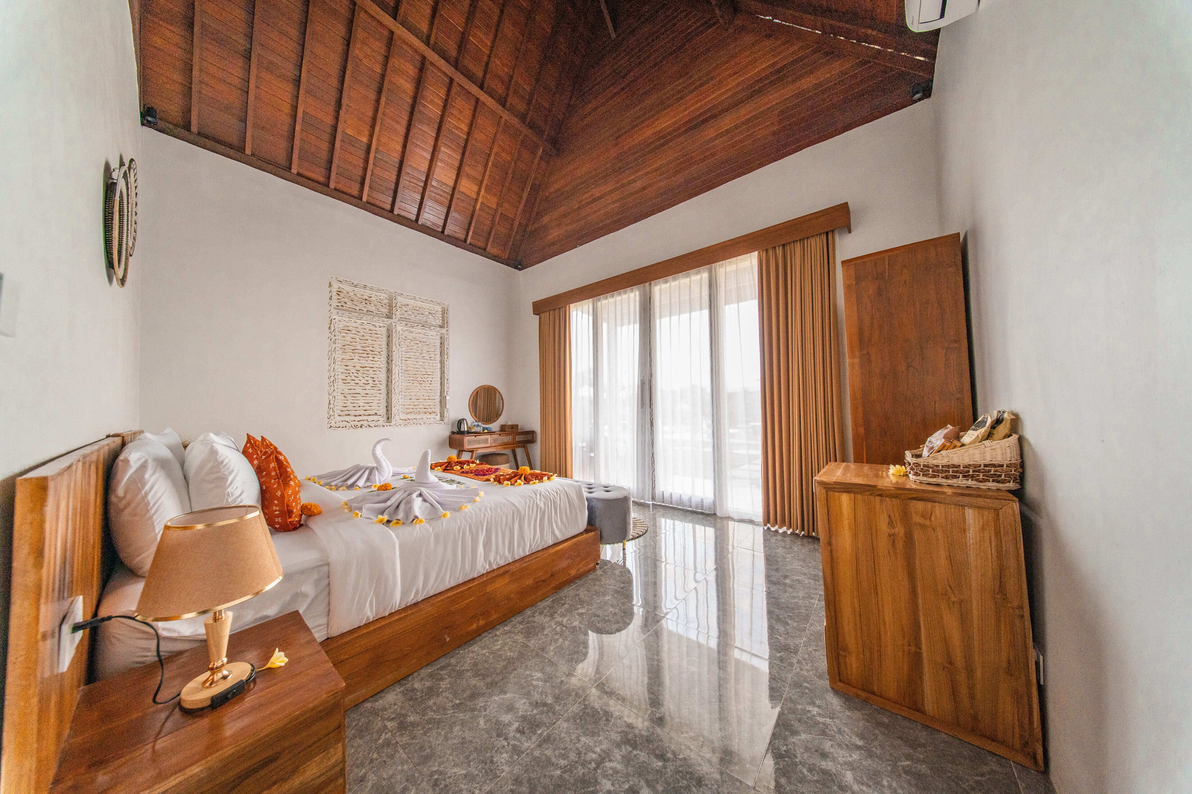 Un dormitorio con techo de madera, una cama con colcha blanca, una mesita de noche de madera con una lámpara, un armario de madera y un ventanal grande con cortinas.