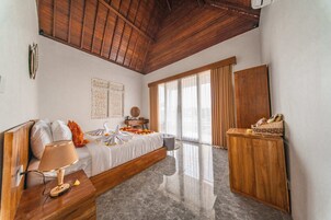 Un dormitorio con techo de madera, una cama con colcha blanca, una mesita de noche de madera con una lámpara, un armario de madera y un ventanal grande con cortinas.