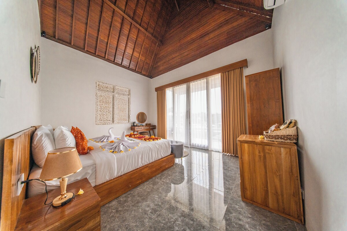 Habitación de hotel con techo de madera, cama con ropa blanca, mesita de noche con lámpara, armario de madera y una ventana grande con cortinas.