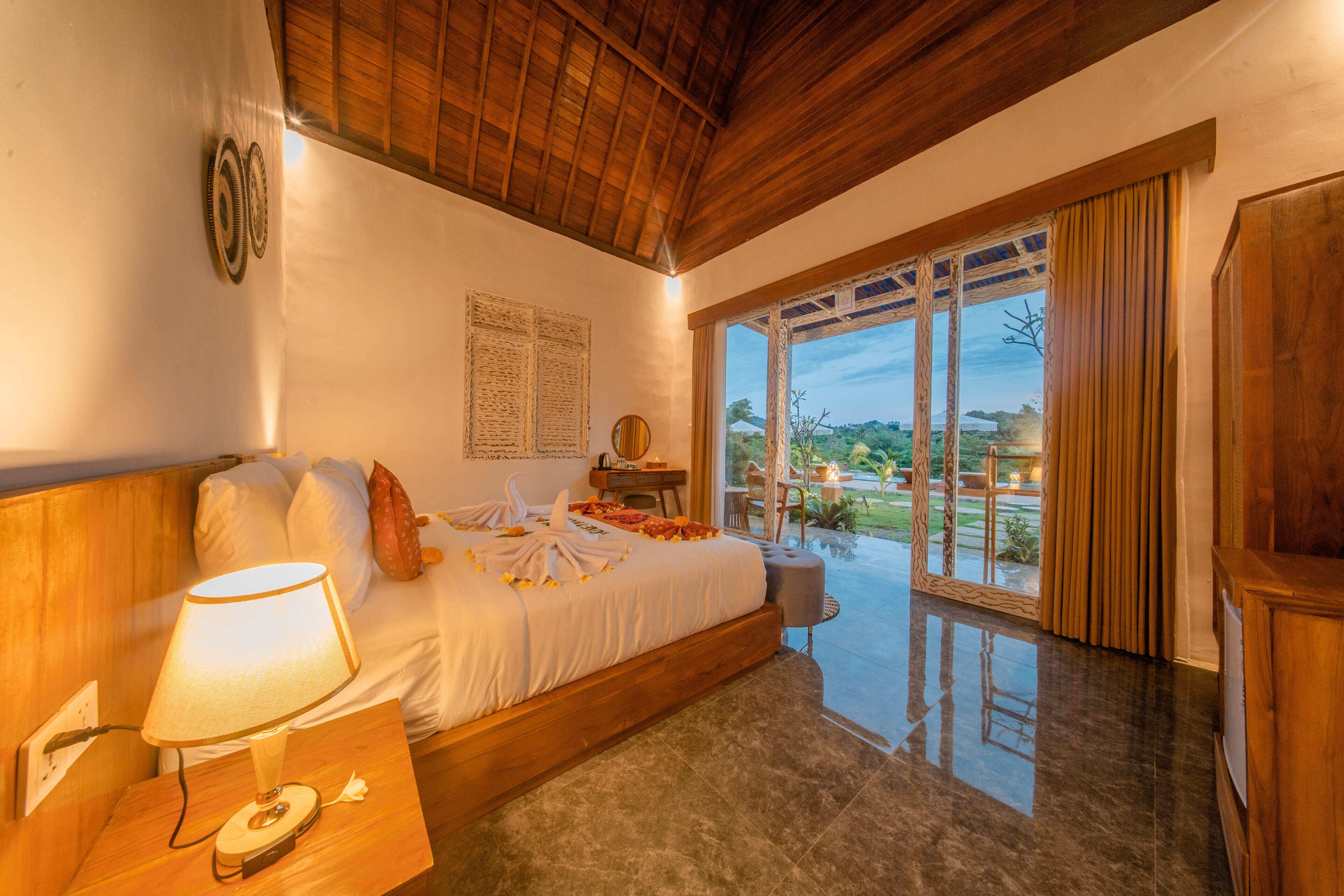 A bedroom with a bed, a bedside lamp, a wooden headboard, a window with curtains, and a view of the outdoors.