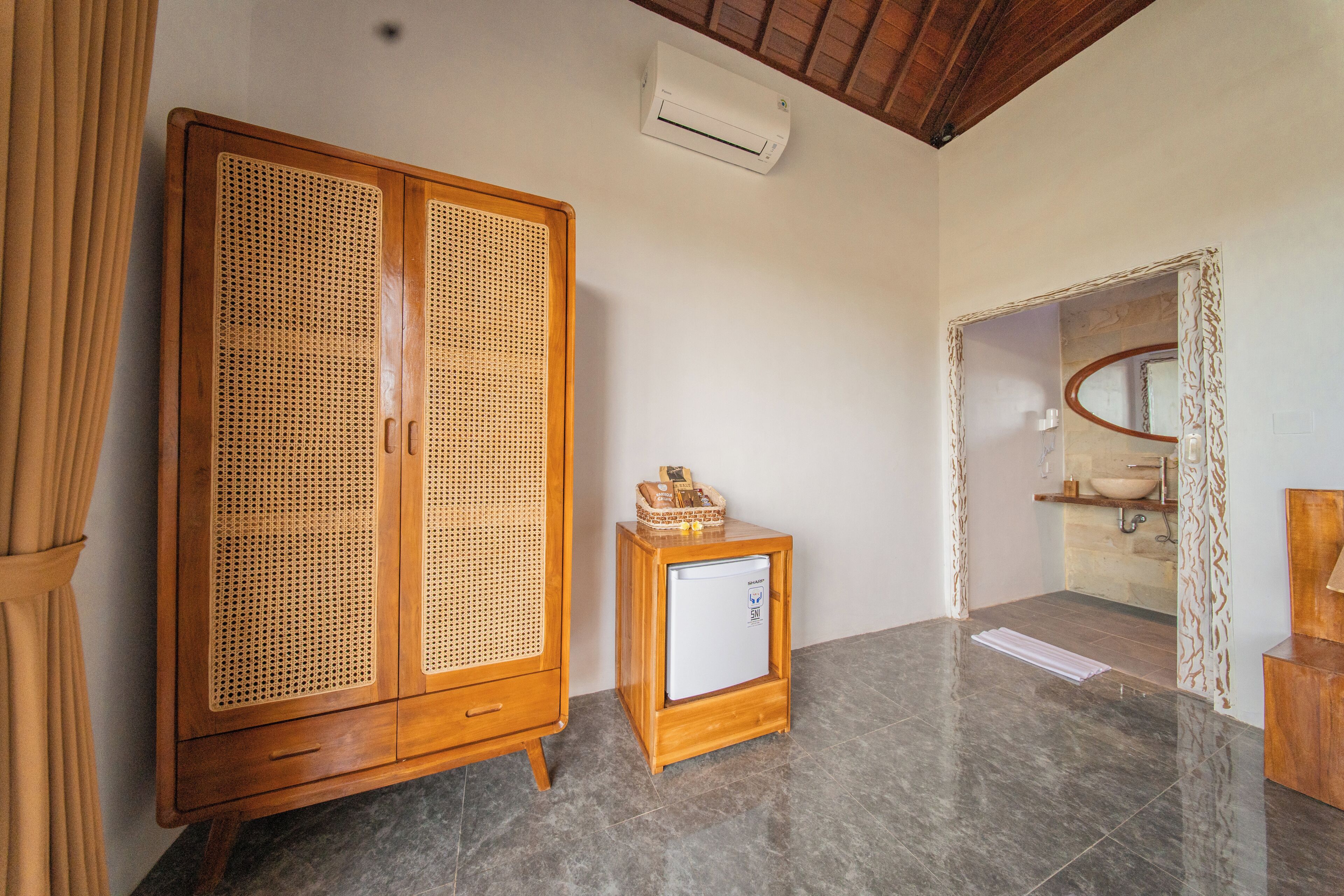 A room with a wooden wardrobe, a small refrigerator, a bathroom with a sink and mirror, and a ceiling fan.