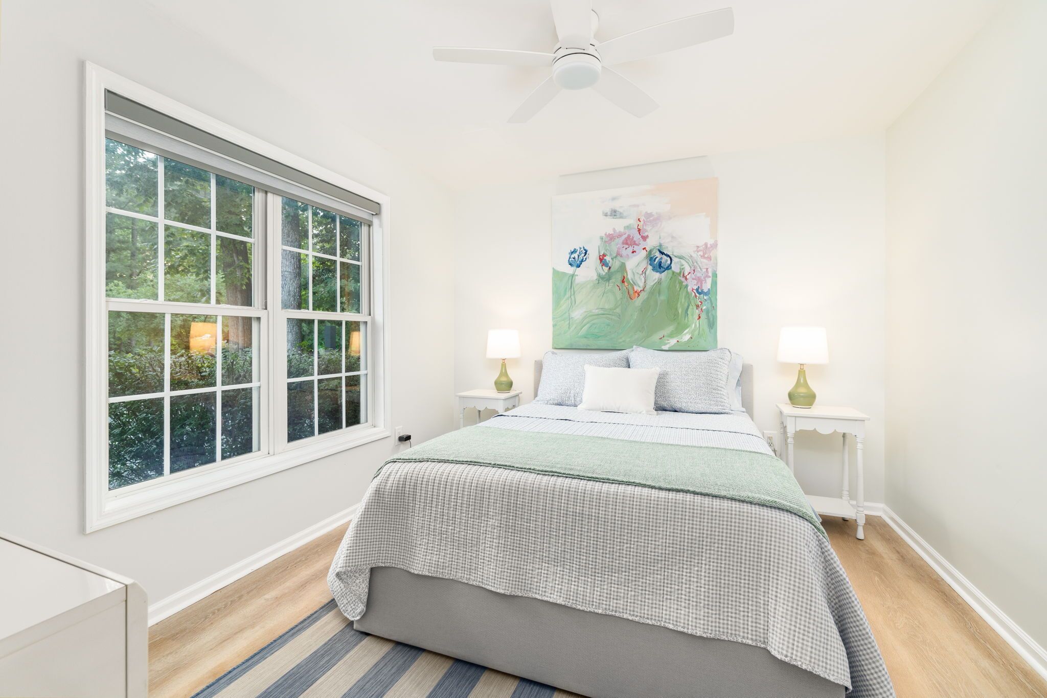 A bedroom with a large window, a ceiling fan, a bed with two bedside tables, and a painting on the wall.