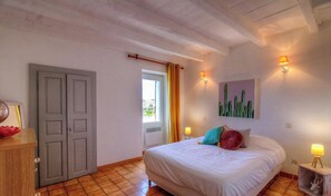 Ein Schlafzimmer mit einem Bett, zwei Nachttischlampen, einer Tür, einem Fenster mit Vorhängen und einem Kakteenbild an der Wand.