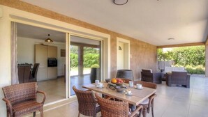 Un comedor con mobiliario de mimbre, una mesa dispuesta con tazas y un frutero, y vistas a una cocina y follaje exterior.