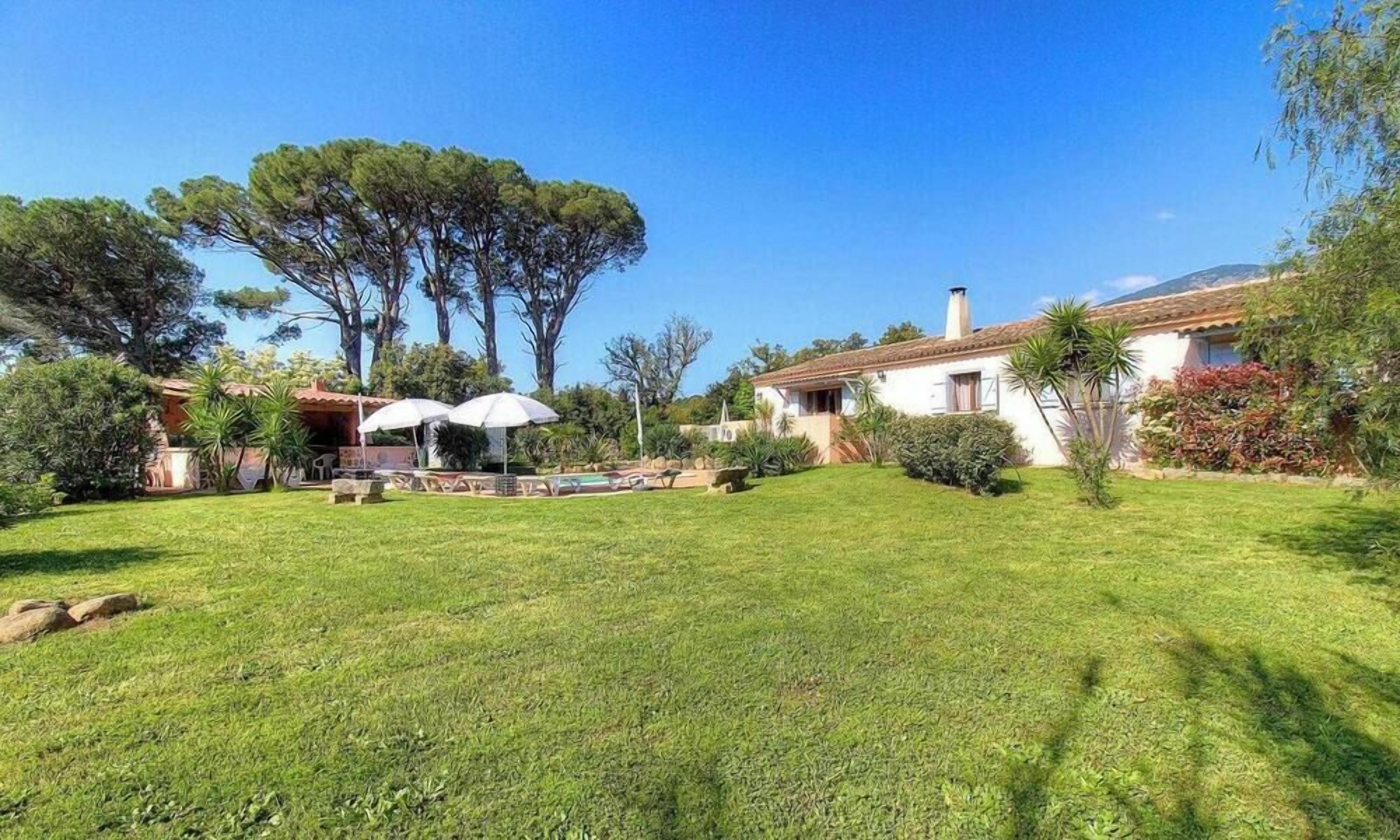 Un jardin bien entretenu avec une piscine, une terrasse agrémentée de parasols et une maison avec un toit en tuiles.