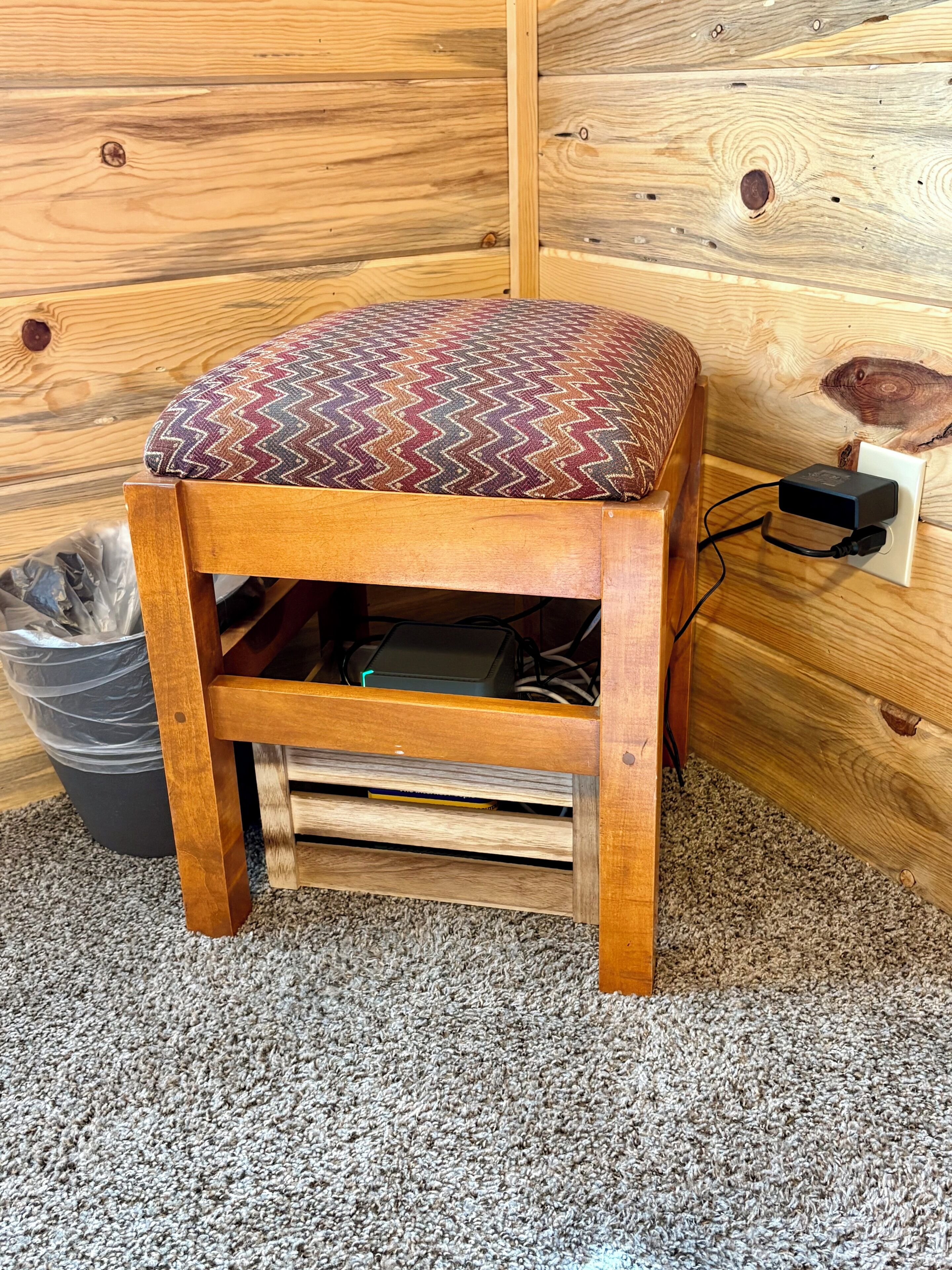 A wooden stool with a patterned cushion, a trash bin, and a power outlet on a carpeted floor.
