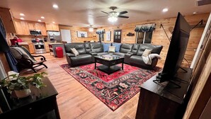 A living room with a black leather sofa set, a coffee table, a red patterned rug, a ceiling fan, wooden walls, and a kitchen area in the background. - B & T's Garden Getaway-Located in Historic Parowan near Brianhead Ski Resort (Parowan)