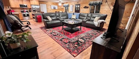 A living room with a black leather sofa set, a coffee table, a red patterned rug, a ceiling fan, wooden walls, and a kitchen area in the background.