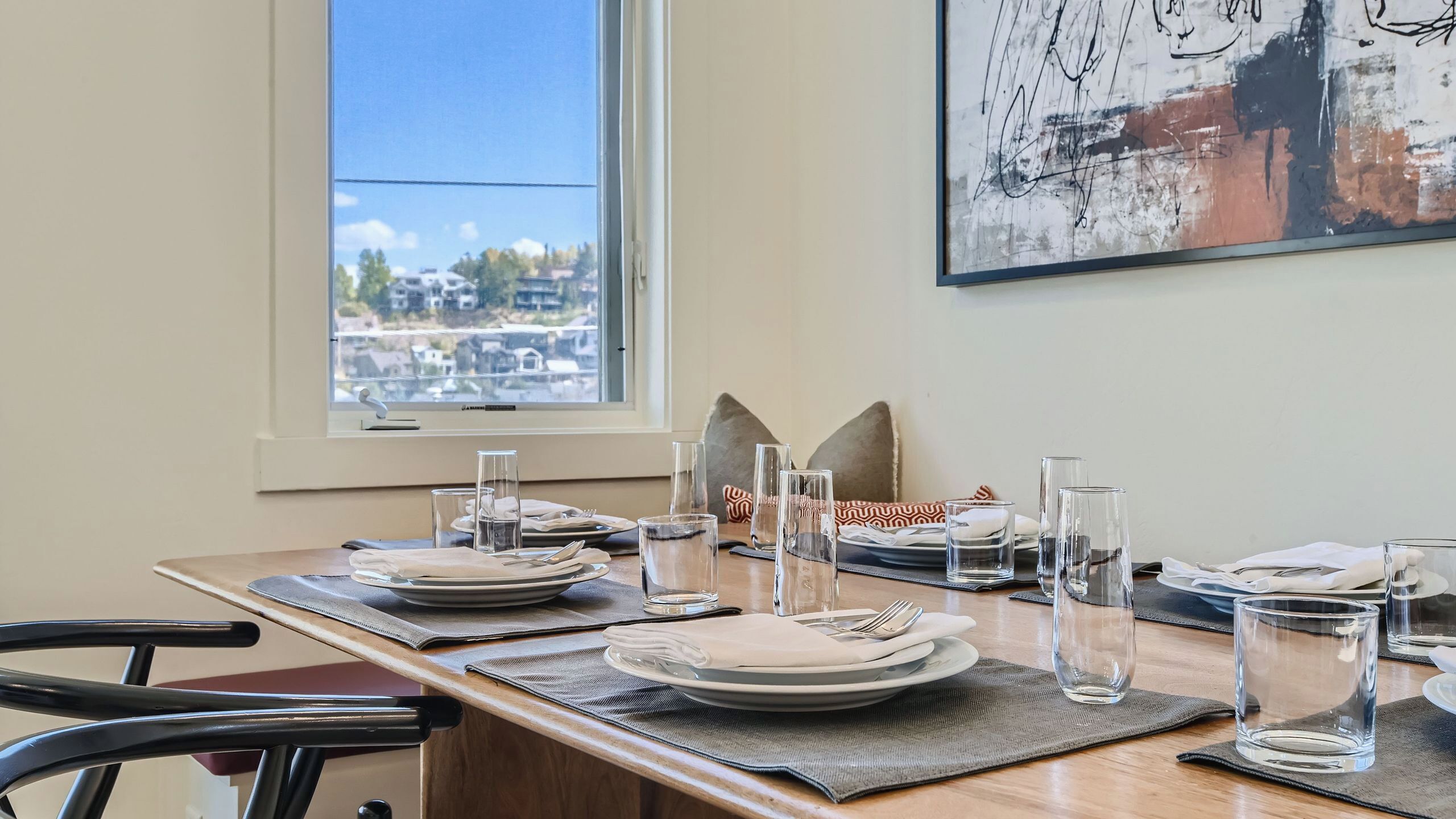 A dining table set with plates, glasses, and cutlery, a window with a view of buildings, and a piece of abstract art on the wall.
