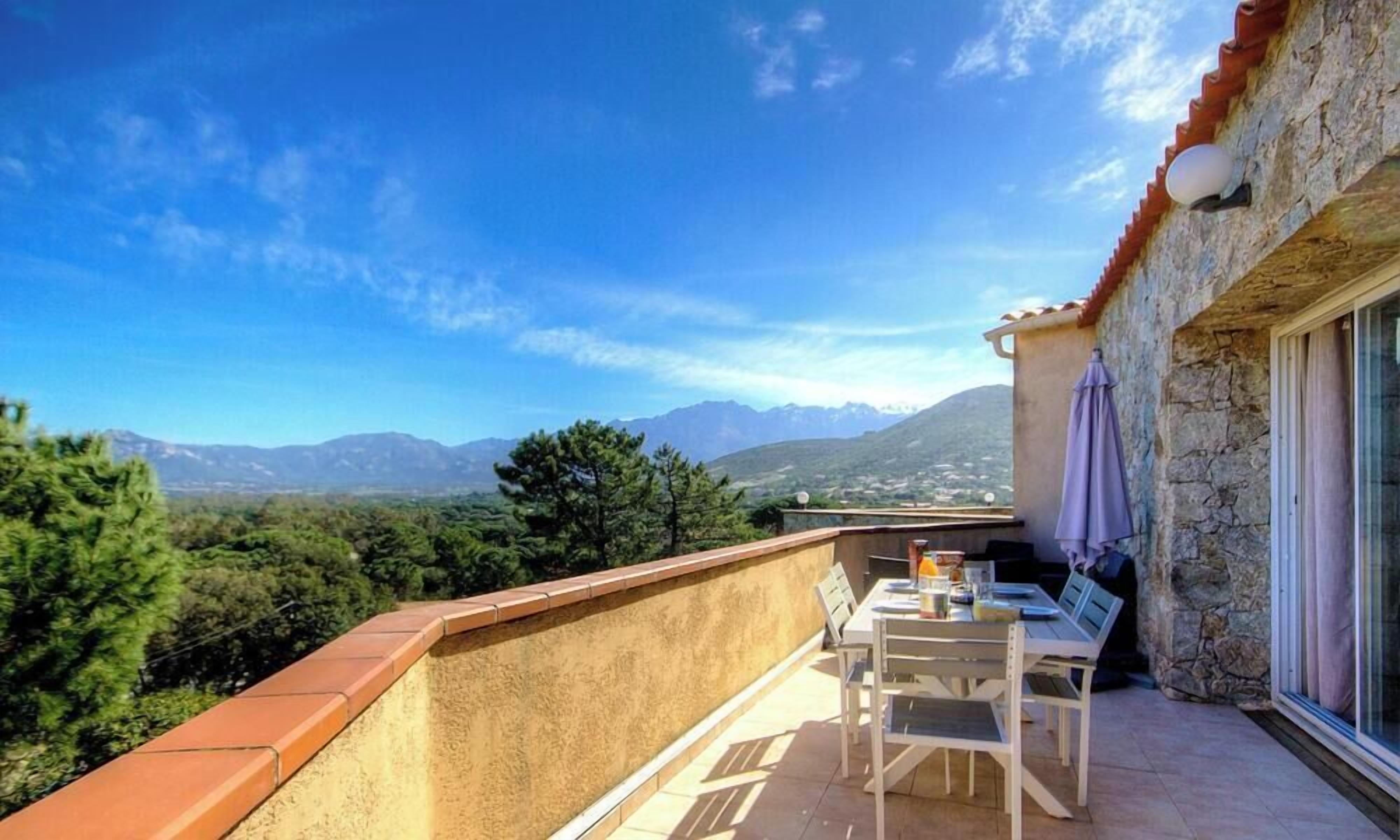 A balcony with a table and chairs, a view of mountains, and a stone wall.