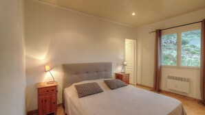 Un dormitorio con una cama, dos mesitas de noche, una mesita auxiliar, una ventana con cortinas y una puerta.