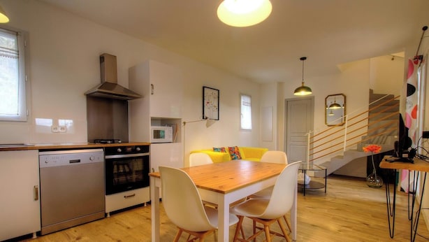Cocina compacta equipada con lavavajillas, horno y microondas. Hay una mesa de comedor de madera con sillas, una escalera y una pequeña zona de estar con un sofá amarillo.