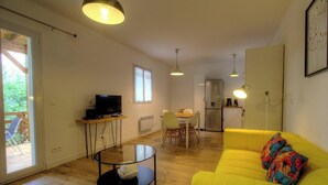 Una sala de estar moderna con un sofá amarillo, una mesa de centro de vidrio y un televisor sobre un soporte de madera. Hay un comedor con mesa y sillas, y una cocina al fondo.
