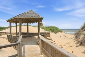 Ein hölzerner Strandpavillon mit geneigtem Dach, ein Holzsteg, der dorthin führt, und sandige Dünen im Hintergrund.