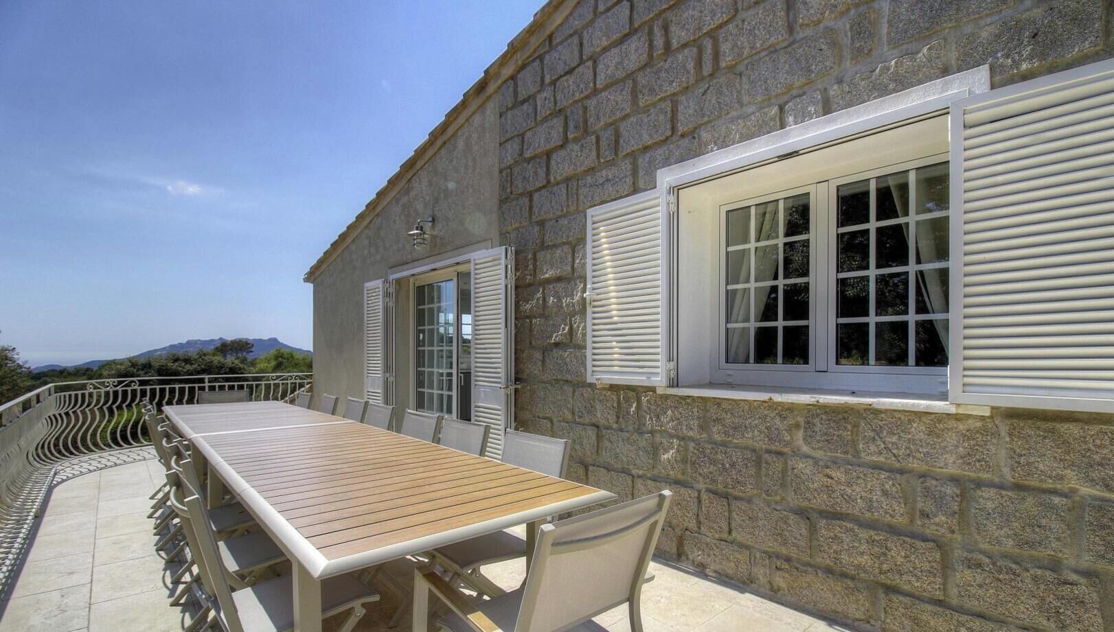 A stone building with a wooden table and chairs outside, a window with shutters, and a metal railing.