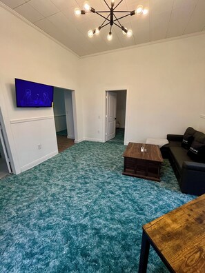 A living room with a blue patterned carpet, a wooden coffee table, a black sofa, a TV mounted on the wall, and a ceiling light fixture.