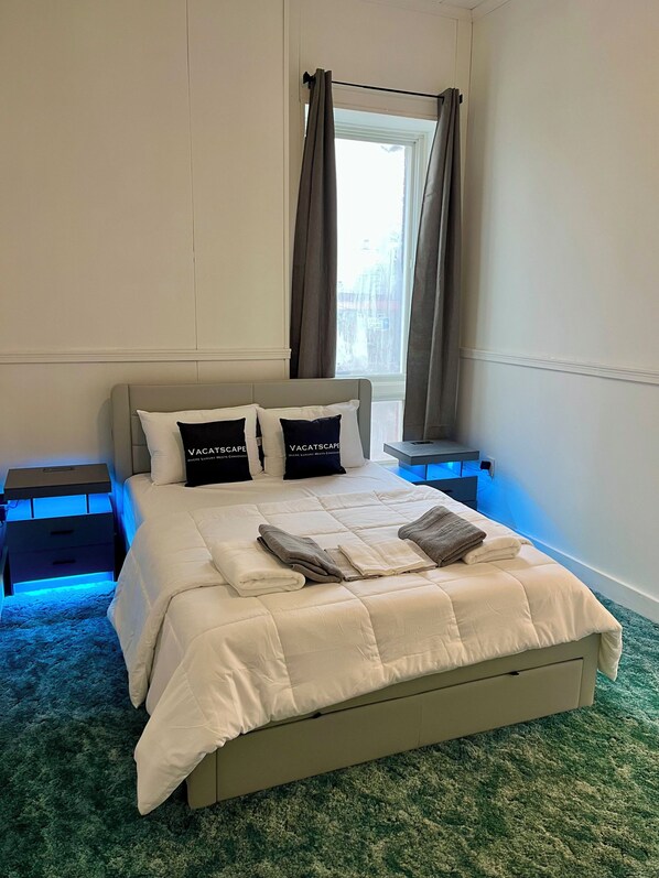 A neatly made bed with pillows, a bedside table, a nightstand, and a window with curtains.