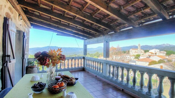Un balcone con un tavolo apparecchiato per mangiare, un vaso con fiori e vista su una città e montagne.