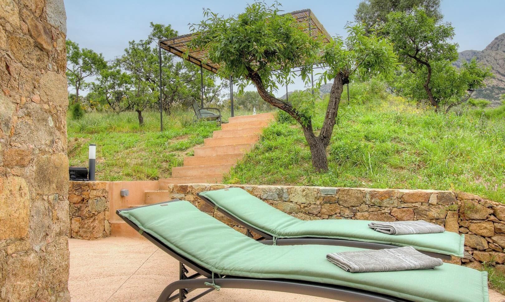 Een stenen muur met een metalen hekwerk, twee groene fauteuils met kussens, en een trap die naar een gebied met bomen leidt.