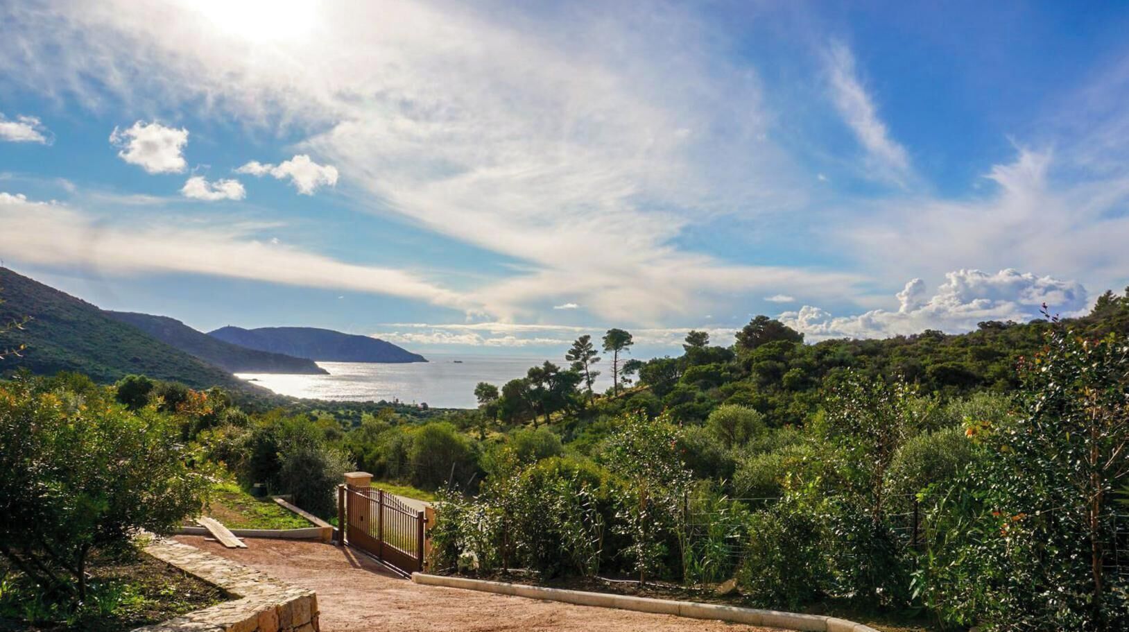 A coastal landscape with a paved path leading to a gate, surrounded by lush greenery and a view of the sea.