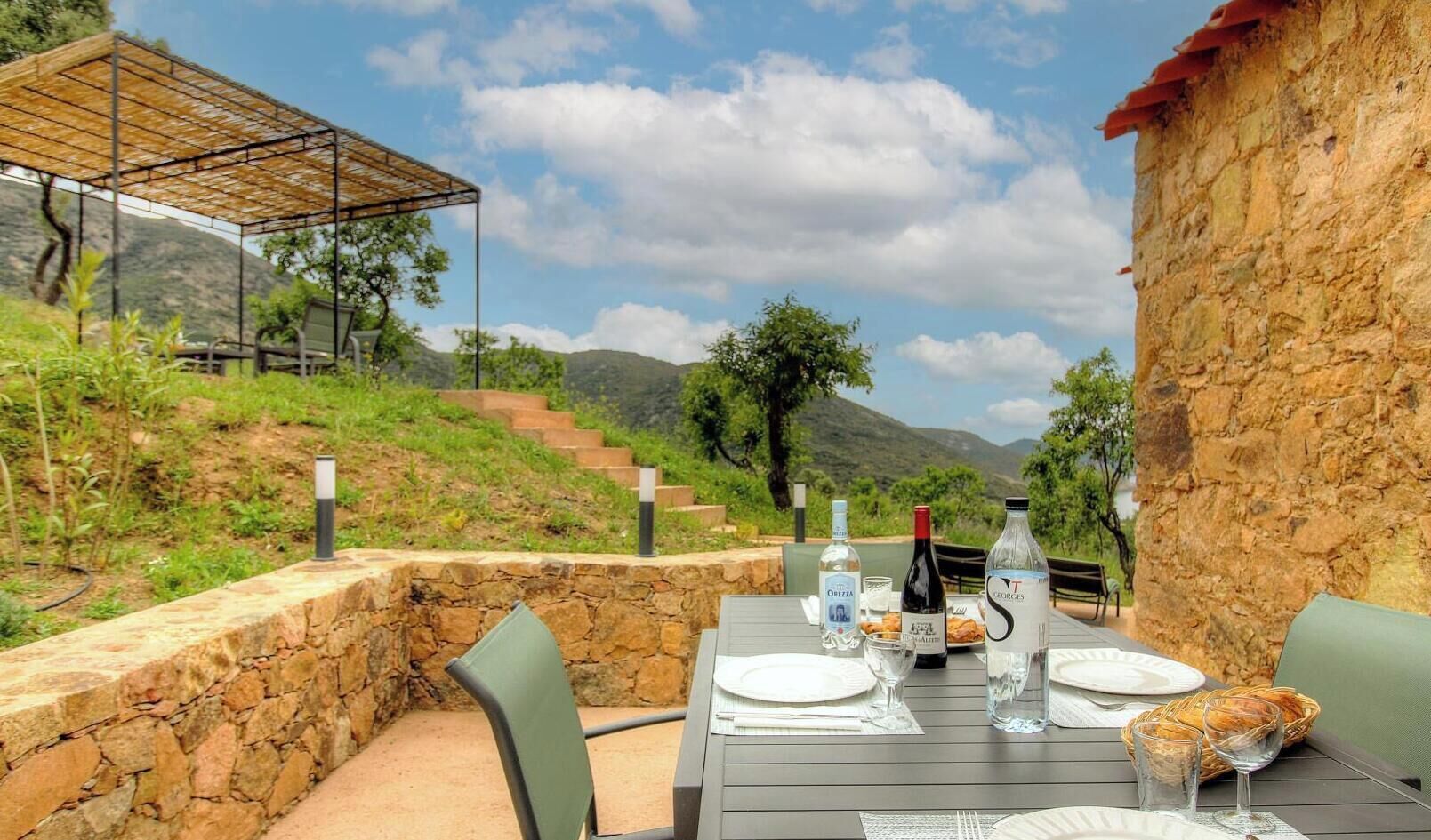 Mesa de comedor al aire libre con pared de piedra, pérgola y montañas al fondo.