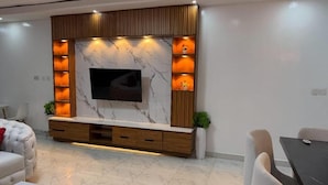A modern living room with a TV mounted on a marble wall, wooden shelves, and a dining area with a table and chairs. - Kigali Savannah Homes and E6 Apartment  (Kigali)