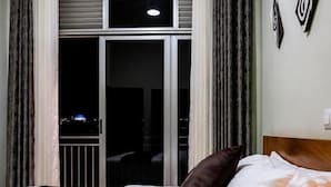 A hotel room with a bed, a nightstand, a lamp, a window with curtains, and a view of the outside. - Kigali Savannah Homes and E6 Apartment  (Kigali)