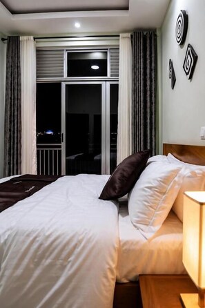 A hotel room with a bed, a nightstand, a lamp, a window with curtains, and a view of the outside.