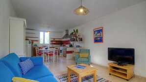 A living room with a blue sofa, a wooden coffee table, a TV on a stand, a kitchen in the background, and a dining area with chairs. - PAMPASGIOLU - PRUNELLI DI FIUM'ORBO (PRUNELLI DI FIUM'ORBO)